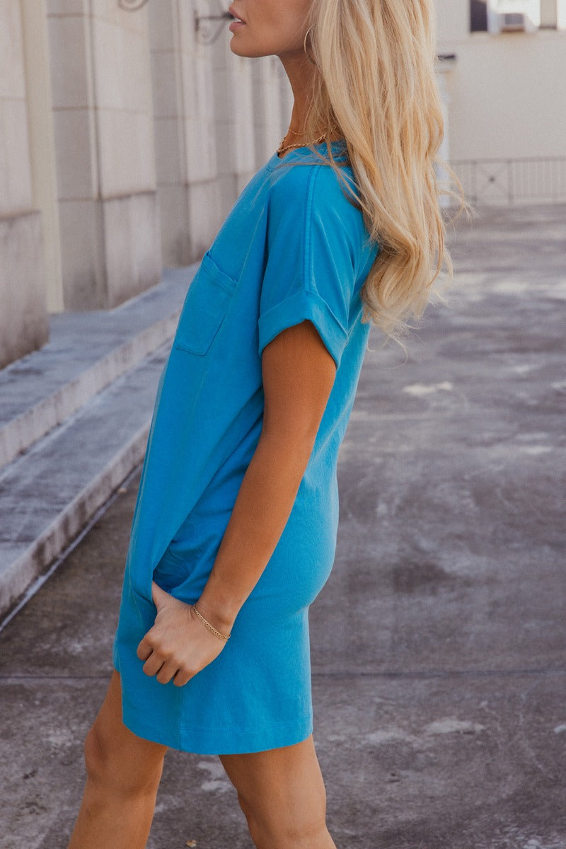 Side view of model wearing the Skylar Blue Short Sleeve Mini Dress that has ocean blue cotton fabric, mini length, two side pockets, a left chest pocket, a round neck, and short sleeves with folded hems.