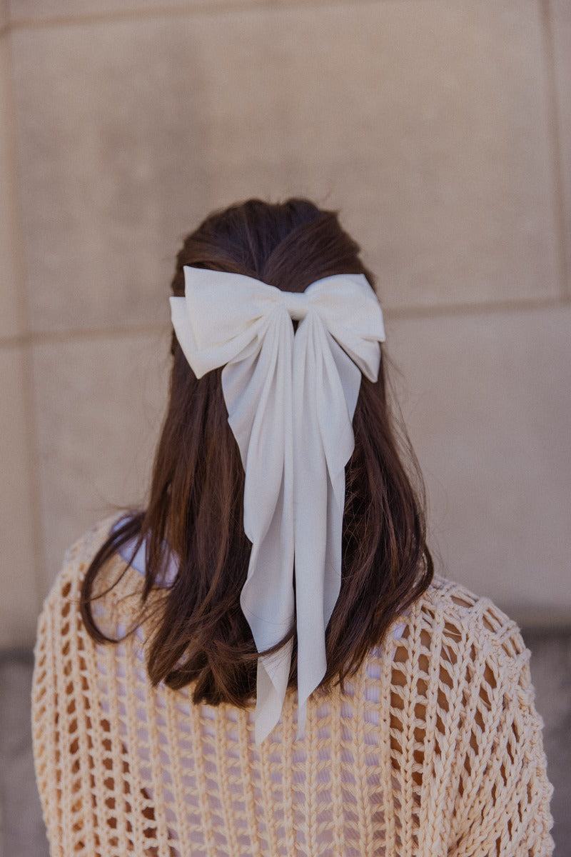 Image shows back of model's head while wearing the Cora Off White Satin Long Hair Bow that features long off white satin fabric shaped as a bow and a clip closure.