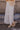Side view of model wearing the Athena White Tiered Maxi Skirt that features white lightweight fabric, a two tiered body with ruffle details, a lace hem, midi length, white lining, and an elastic waistband.