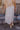 Back view of model wearing the Athena White Tiered Maxi Skirt that features white lightweight fabric, a two tiered body with ruffle details, a lace hem, midi length, white lining, and an elastic waistband.