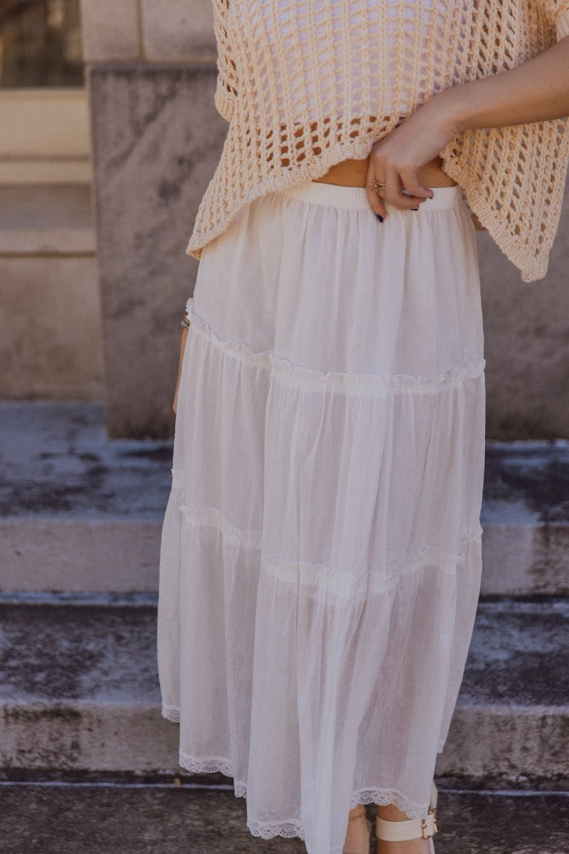 front view of model wearing the Athena White Tiered Maxi Skirt that features white lightweight fabric, a two tiered body with ruffle details, a lace hem, midi length, white lining, and an elastic waistband.