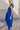 Side view of model wearing the Kristina Royal Blue Smocked Sleeveless Midi Dress that has royal blue fabric, side slit pockets, midi length, smocked upper, a scoop neckline, and ruffled straps.