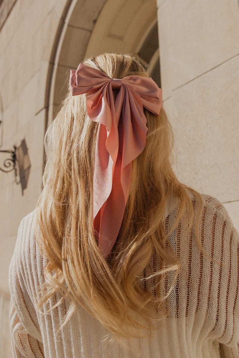Image shows back of model's head while wearing the Cora Pink Satin Long Hair Bow that has long blush pink satin fabric shaped as a bow and a clip closure.