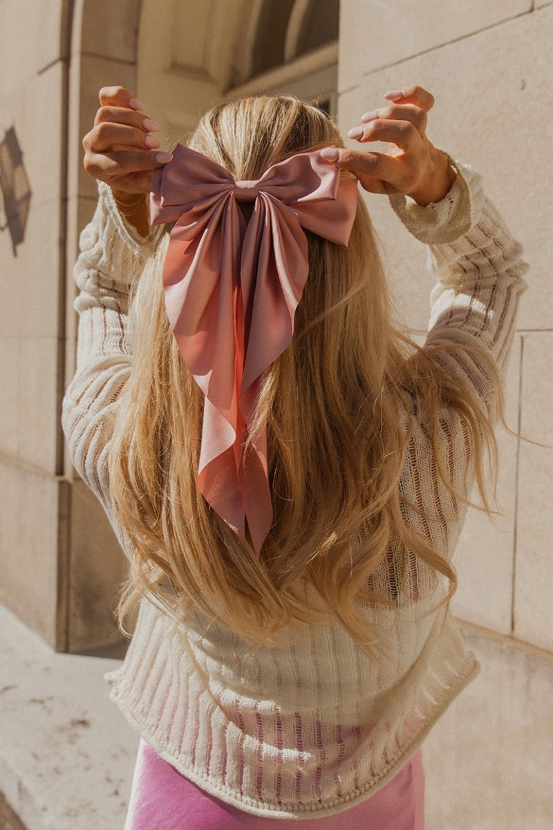 Image shows back of model's head while wearing the Cora Pink Satin Long Hair Bow that has long blush pink satin fabric shaped as a bow and a clip closure.