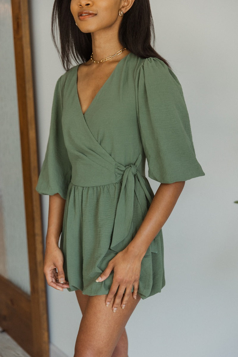 close front view of model wearing the Celia Romper in Green that has green lightweight fabric, a bubble hem, a tie around the waist, a surplice neck, half puff sleeves with elastic trim, and a back zipper with a hook closure