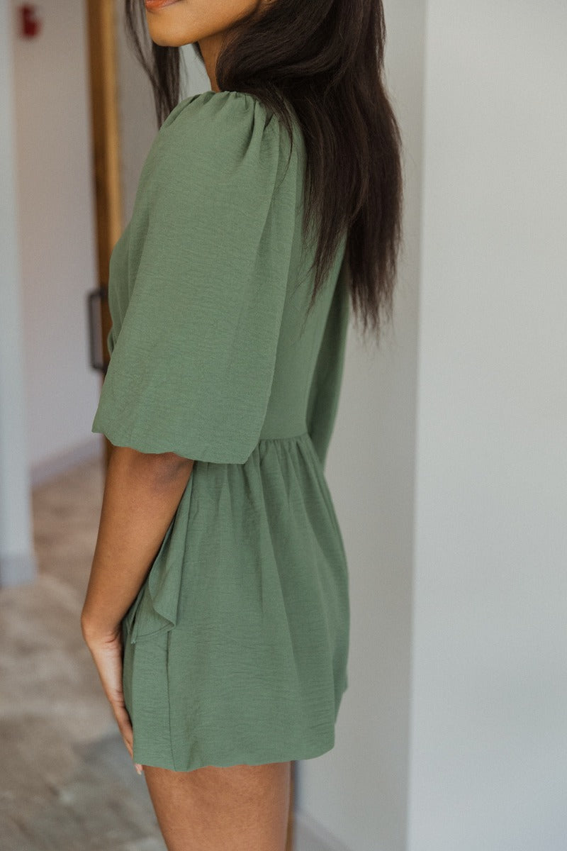 close side view of model wearing the Celia Romper in Green that has green lightweight fabric, a bubble hem, a tie around the waist, a surplice neck, half puff sleeves with elastic trim, and a back zipper with a hook closure
