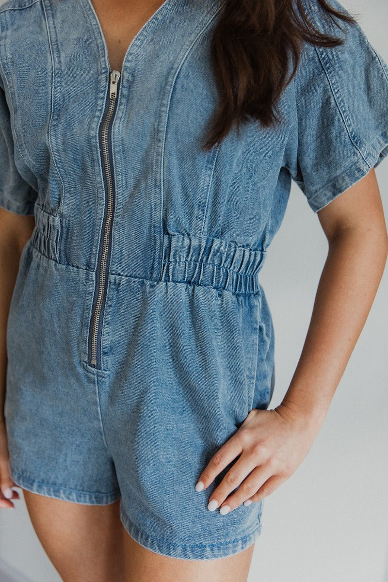 Close-up front view of model wearing the Denim Dreams Romper that has light wash denim fabric, two front pockets, an elastic waistband, a front zipper closure, a plunge neckline and short sleeves