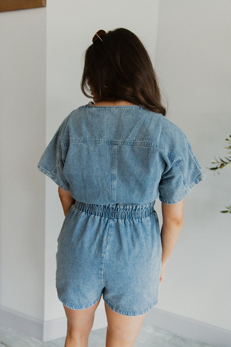 Back view of model wearing the Denim Dreams Romper that has light wash denim fabric, two front pockets, an elastic waistband, a front zipper closure, a plunge neckline and short sleeves