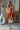 Full body front view of model wearing the Falling For You Dress that has burnt orange fabric, mini length, burnt orange lining, a flare skirt with a tiered ruffle design, a square neckline, thick straps, and a monochromatic back zipper