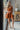 Full body front view of model wearing the Falling For You Dress that has burnt orange fabric, mini length, burnt orange lining, a flare skirt with a tiered ruffle design, a square neckline, thick straps, and a monochromatic back zipper