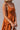 side view of model wearing the Falling For You Dress that has burnt orange fabric, mini length, burnt orange lining, a flare skirt with a tiered ruffle design, a square neckline, thick straps, and a monochromatic back zipper
