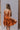 Back view of model wearing the Falling For You Dress that has burnt orange fabric, mini length, burnt orange lining, a flare skirt with a tiered ruffle design, a square neckline, thick straps, and a monochromatic back zipper