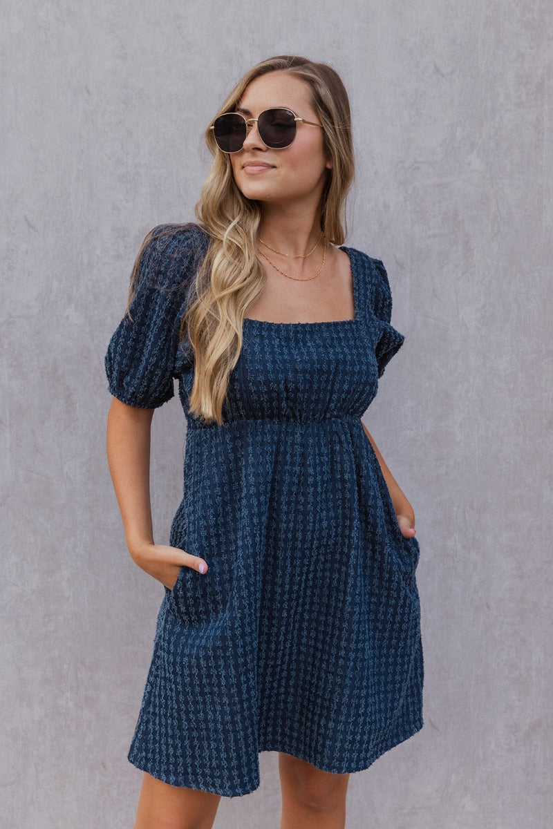 Front view of model wearing the Thinking Of You Dress that has indigo blue denim fabric with a textured design, mini length, an elastic waistband, pockets on each side, a square neckline and short puff sleeves with elastic hems.