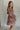 Side view of model wearing the Floral Fantasy Dress that has semi-sheer fabric with a purple, pink, green, red, orange and cream floral design, mini length, a flare skirt, a smocked upper, a square neck with lettuce trim, and long puff sleeves.