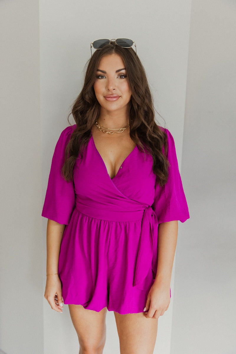 Front view of model wearing the Celia Romper in Magenta which features magenta lightweight fabric, a bubble hem, magenta lining, a tie around the waist, a surplice neckline, half puff sleeves with elastic trim, and a monochromatic back zipper with a hook 