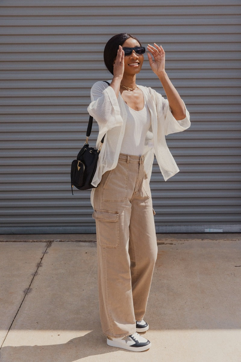 Full body view of model wearing the Ready or Not Cargo Pants in Brown that have light brown denim fabric, cargo pockets on each side, a front zipper with a button closure, two front pockets, belt loops and wide pant legs