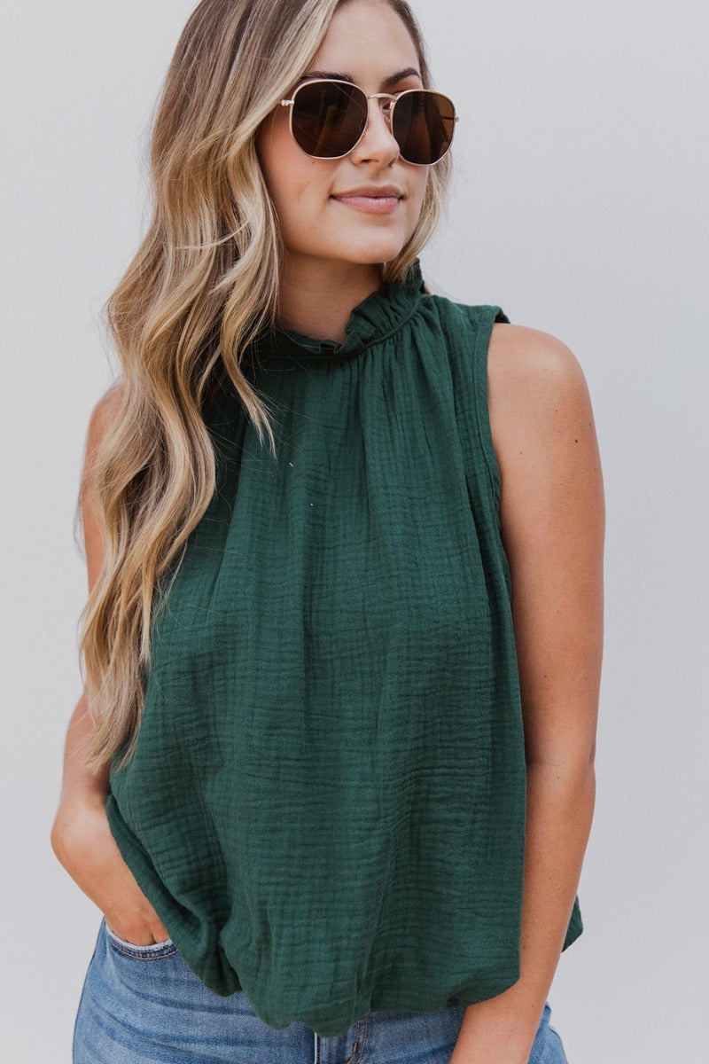 Front view of model wearing the Pretty Please Bubble-Hem Tank that has hunter green gauze fabric, a bubble hem, a halter neckline with ruffle trim, and a sleeveless design.