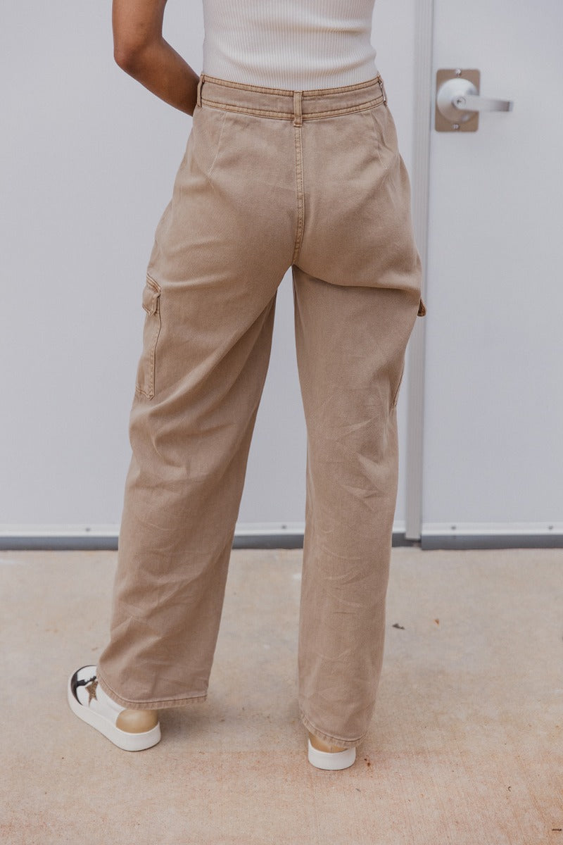 Back view of model wearing the Ready or Not Cargo Pants in Brown that have light brown denim fabric, cargo pockets on each side, a front zipper with a button closure, two front pockets, belt loops and wide pant legs