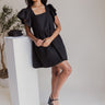 Full body front view of model wearing the Love Is In The Air Dress that has black woven fabric, pockets on each side, a square neckline, and short ruffle sleeves