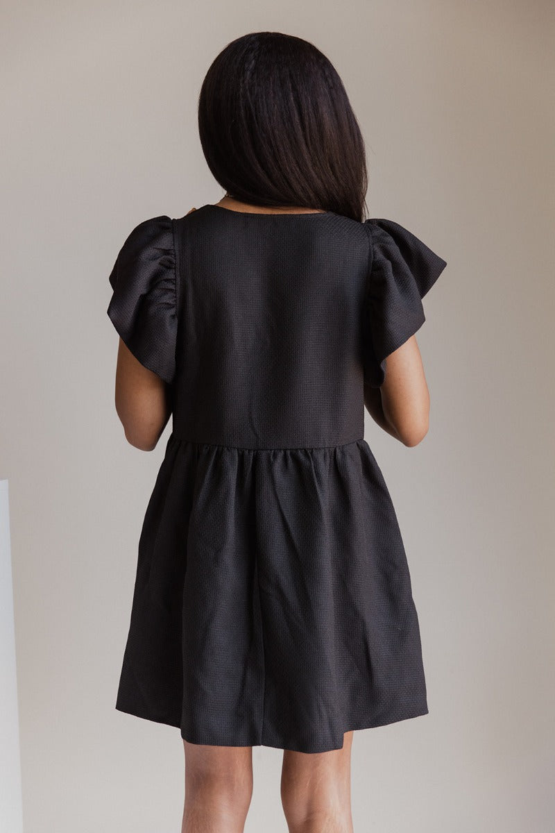 Back view of model wearing the Love Is In The Air Dress that has black woven fabric, pockets on each side, a square neckline, and short ruffle sleeves