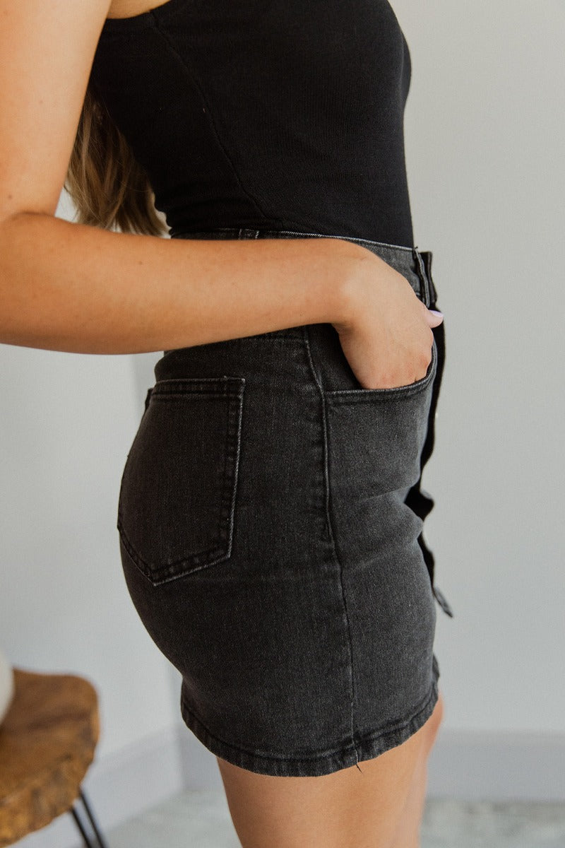 Side view of model wearing the Follow Your Heart Denim Skirt that has black denim fabric, mini length, two front pockets, two back pockets, belt loops, and white heart button-up closures