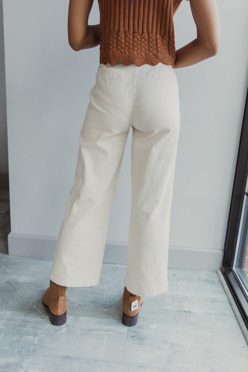 Back view of model wearing the Good Day Pants in Cream that have cream denim fabric, pockets, copper button up closures, belt loops and wide cropped flares.