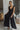 Full body view of model wearing the Moment In Time Jumpsuit in Black which features black lightweight fabric, black lining, wide leg pants, criss cross detail at waist, a plunge neckline, ruffle straps, corset ties in the back, an open back and a monochro