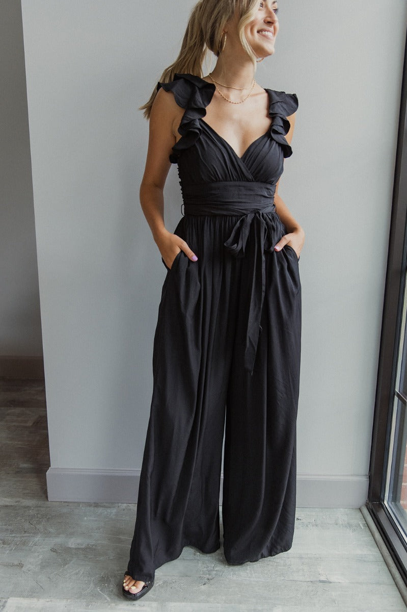 Full body view of model wearing the Moment In Time Jumpsuit in Black which features black lightweight fabric, black lining, wide leg pants, criss cross detail at waist, a plunge neckline, ruffle straps, corset ties in the back, an open back and a monochro