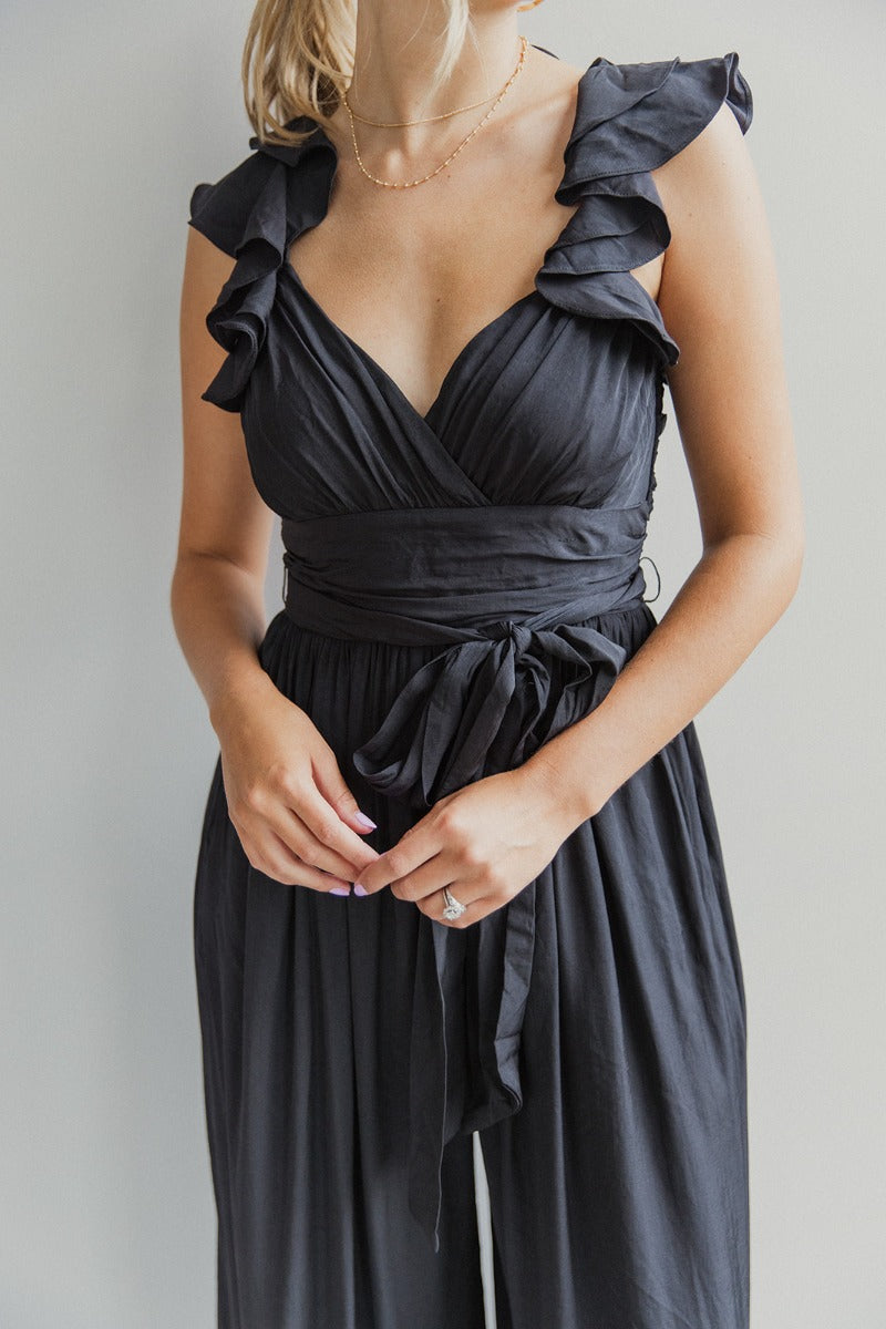 Close up front view of model wearing the Moment In Time Jumpsuit in Black which features black lightweight fabric, black lining, wide leg pants, criss cross detail at waist, a plunge neckline, ruffle straps, corset ties in the back, an open back and a mon