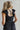 Close up back view of model wearing the Moment In Time Jumpsuit in Black which features black lightweight fabric, black lining, wide leg pants, criss cross detail at waist, a plunge neckline, ruffle straps, corset ties in the back, an open back and a mono