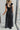 Full body back view of model wearing the Moment In Time Jumpsuit in Black which features black lightweight fabric, black lining, wide leg pants, criss cross detail at waist, a plunge neckline, ruffle straps, corset ties in the back, an open back and a mon