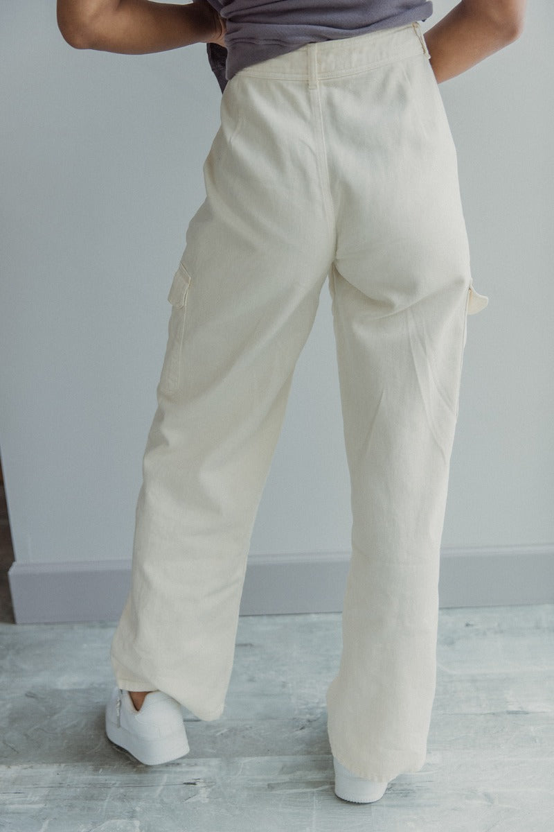 Back view of model wearing the Charmed Life Cargo Pants in Cream which features cream cotton fabric, cargo pockets on each side, a front zipper with a button closure, two front pockets, belt loops and wide pant legs.
