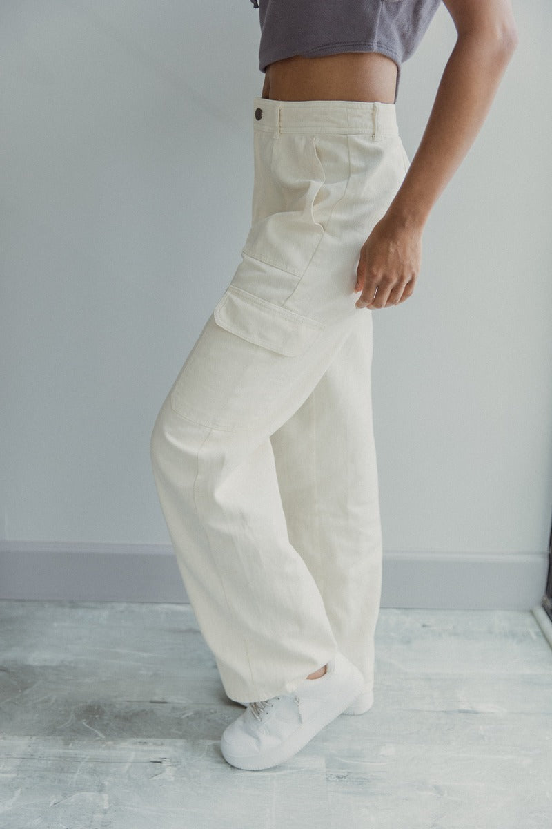Side view of model wearing the Charmed Life Cargo Pants in Cream which features cream cotton fabric, cargo pockets on each side, a front zipper with a button closure, two front pockets, belt loops and wide pant legs.