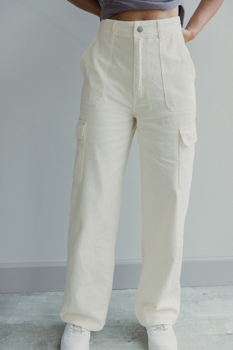 Front view of model wearing the Charmed Life Cargo Pants in Cream which features cream cotton fabric, cargo pockets on each side, a front zipper with a button closure, two front pockets, belt loops and wide pant legs.