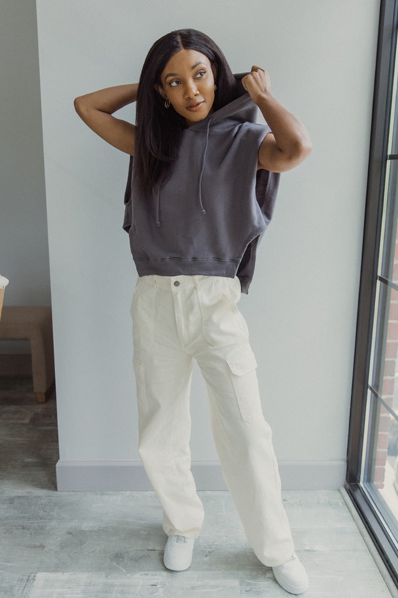 Full body view of model wearing the Charmed Life Cargo Pants in Cream which features cream cotton fabric, cargo pockets on each side, a front zipper with a button closure, two front pockets, belt loops and wide pant legs.