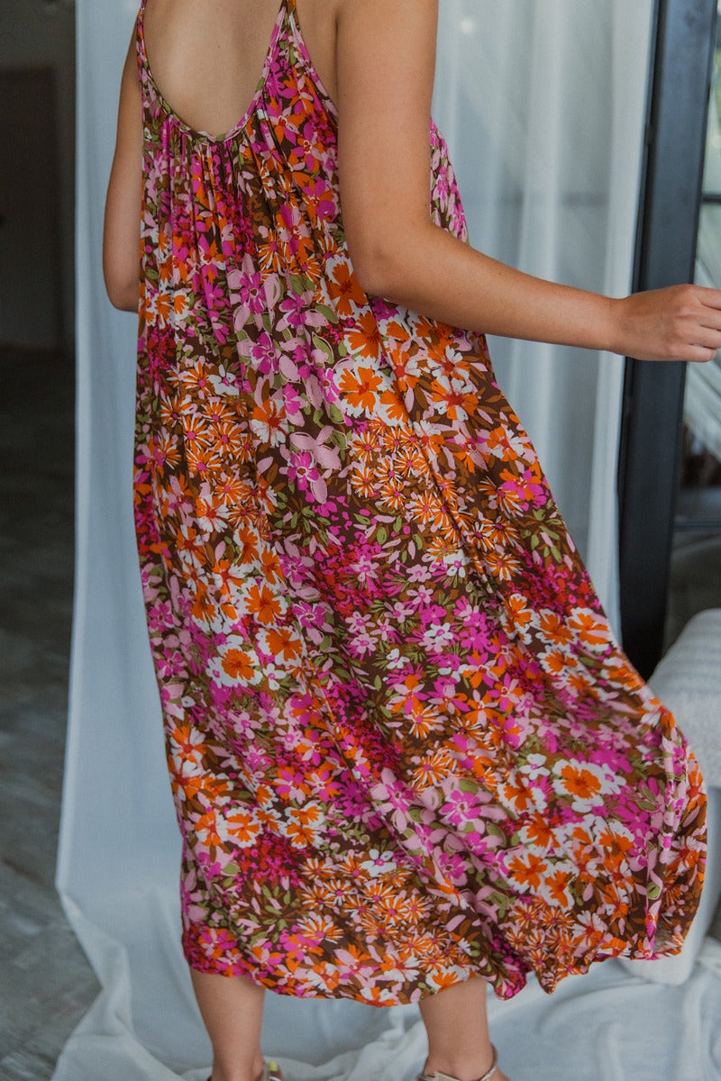 Back view of model wearing the Everything To Me Floral Dress which features a brown, orange, pink, green and white floral print, midi length, a scooped neckline, and adjustable straps.