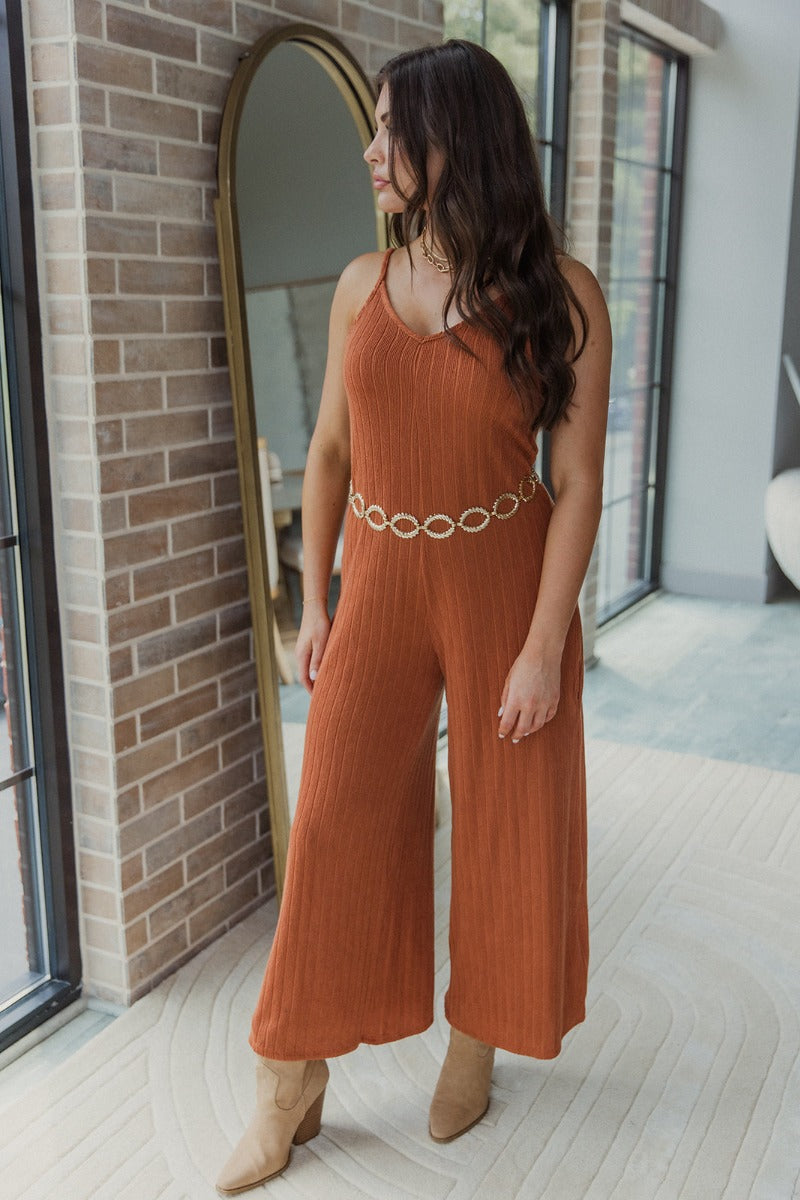 Full body view of model wearing the Unexpected Treasure Jumpsuit which features rust ribbed fabric, wide cropped pants, pockets on each side, a v-neckline, and spaghetti straps.
