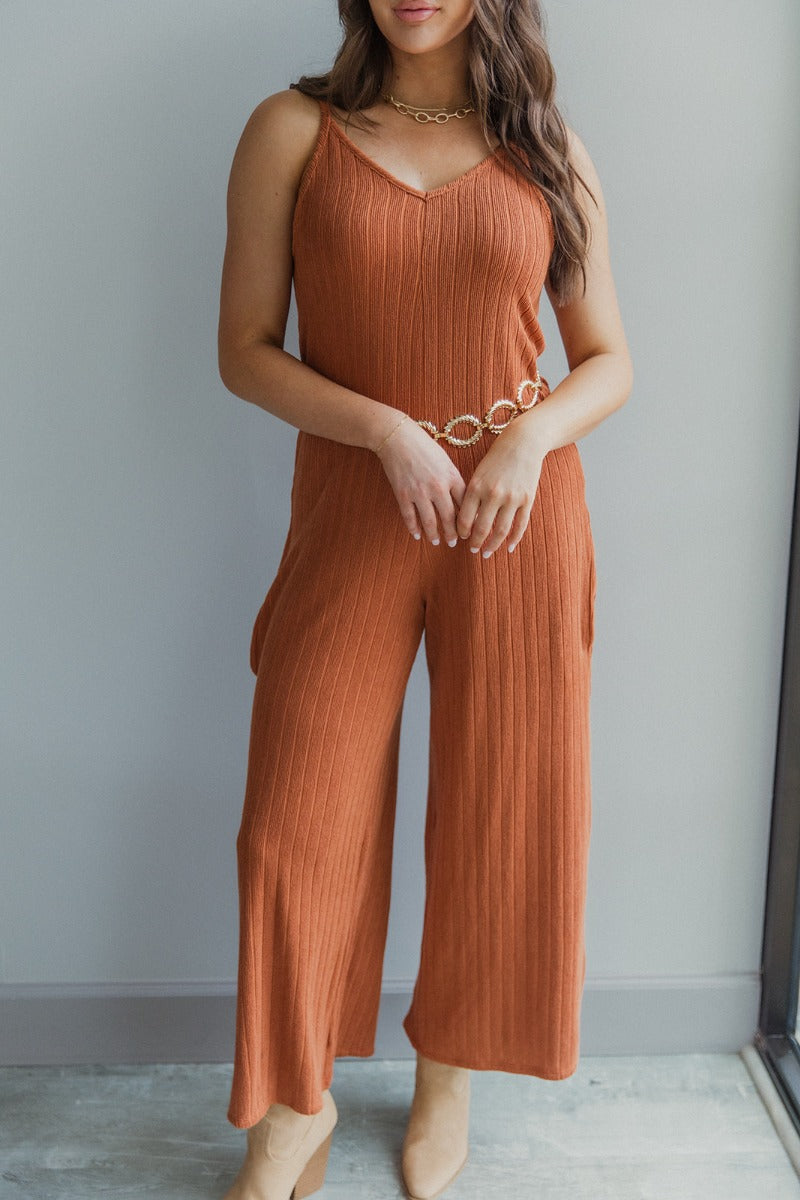Full body view of model wearing the Unexpected Treasure Jumpsuit which features rust ribbed fabric, wide cropped pants, pockets on each side, a v-neckline, and spaghetti straps.