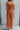 Full body back view of model wearing the Unexpected Treasure Jumpsuit which features rust ribbed fabric, wide cropped pants, pockets on each side, a v-neckline, and spaghetti straps.