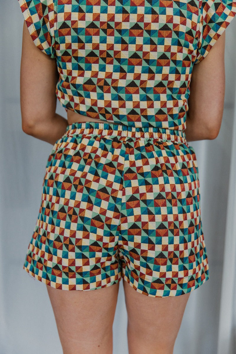 Back view of model wearing the Take Me Places Short which features maroon, rust, yellow, mint, teal, black and cream fabric with a geometric pattern, two front pockets, and an elastic waistband.