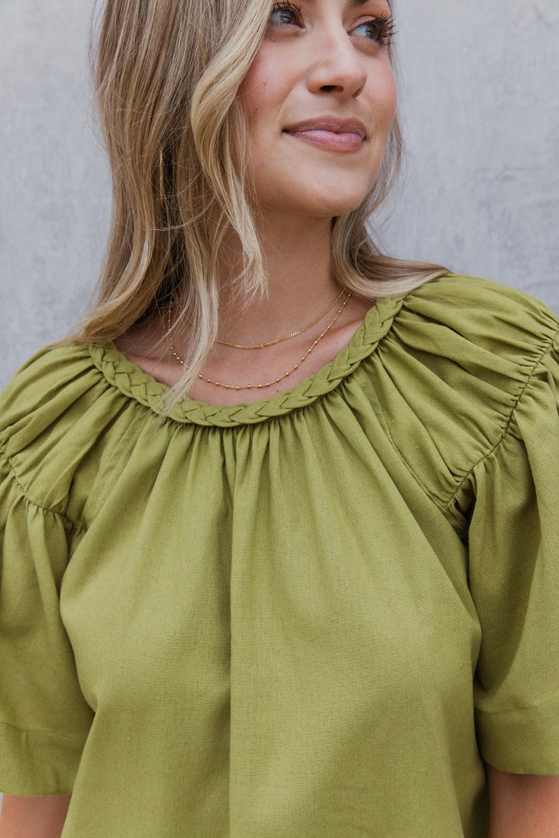 Close up view of model wearing the Stay Kind Top which features olive green fabric, a round neckline with braided details, short flare sleeves and a back keyhole with a button closure.