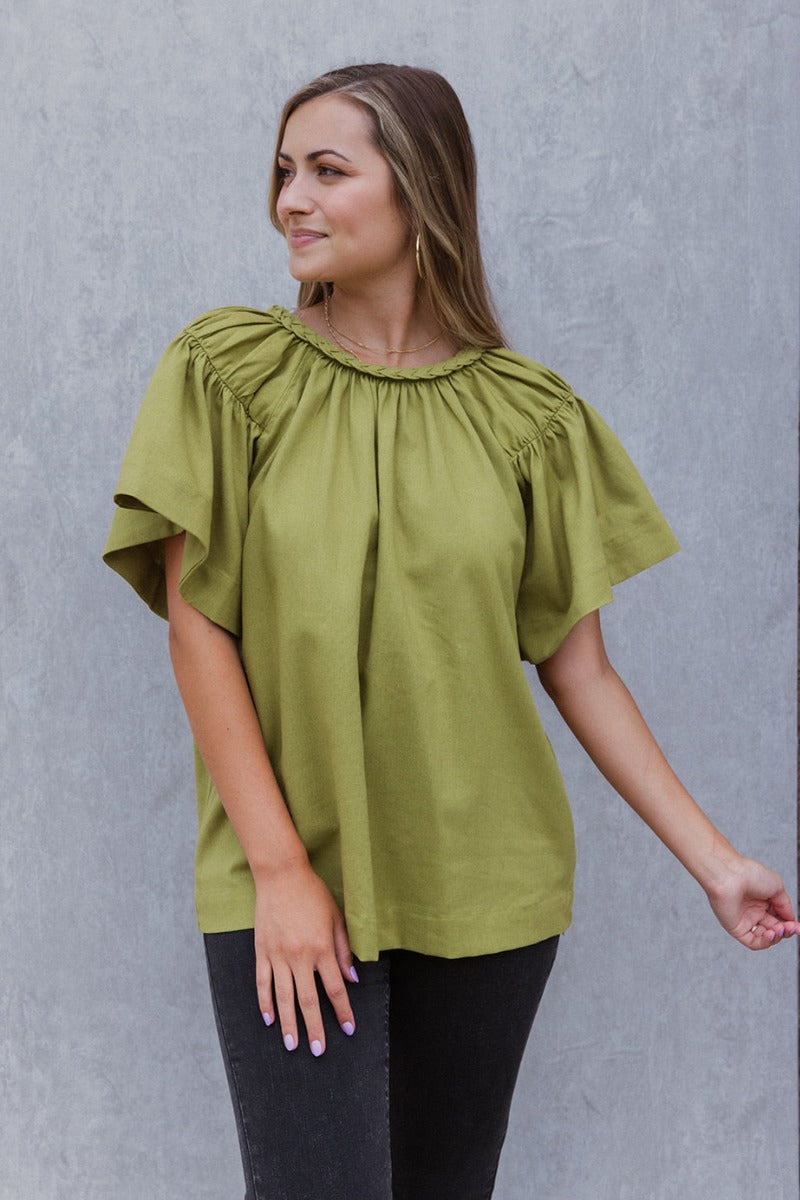 Front view of model wearing the Stay Kind Top which features olive green fabric, a round neckline with braided details, short flare sleeves and a back keyhole with a button closure.
