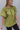 Frontal side view of model wearing the Stay Kind Top which features olive green fabric, a round neckline with braided details, short flare sleeves and a back keyhole with a button closure.