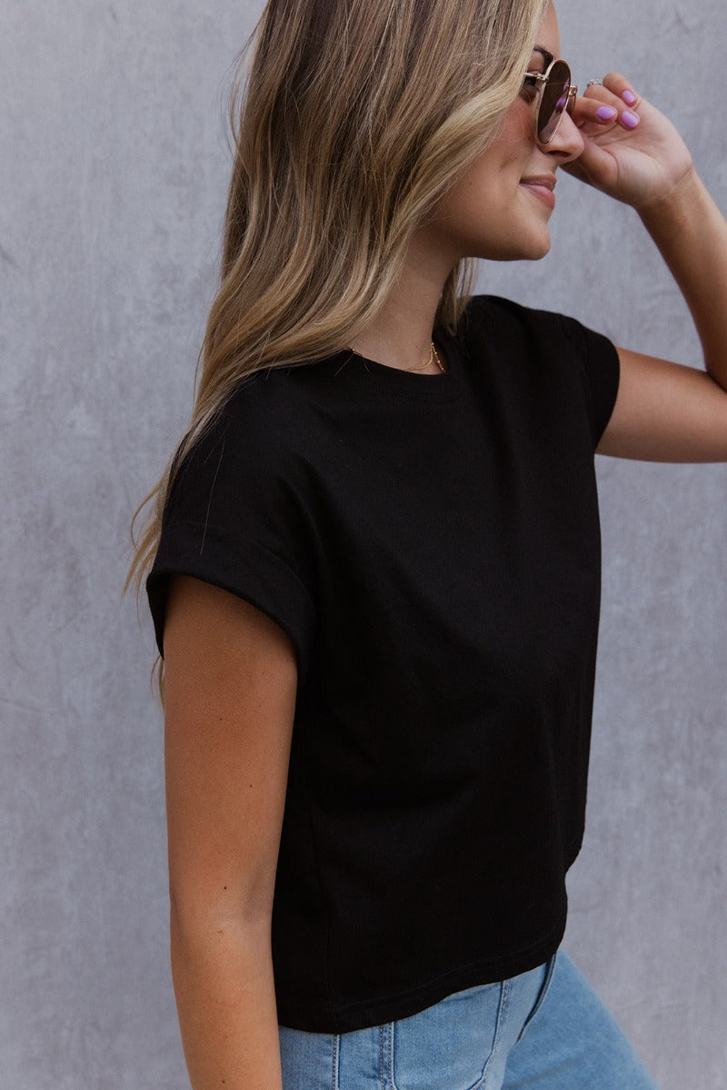 Side view of model wearing the See You Later Tee that has black cotton fabric, a cropped waist, a round neckline and short folded sleeves