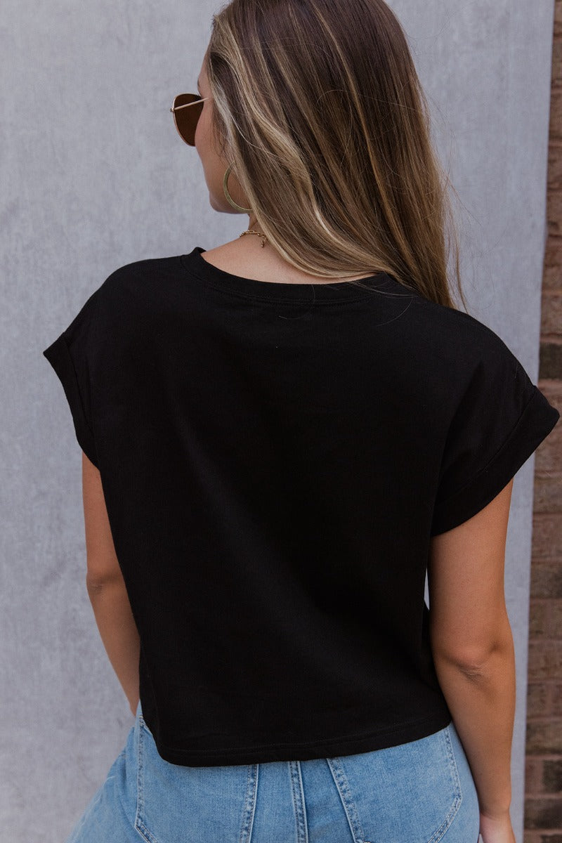 Back view of model wearing the See You Later Tee that has black cotton fabric, a cropped waist, a round neckline and short folded sleeves