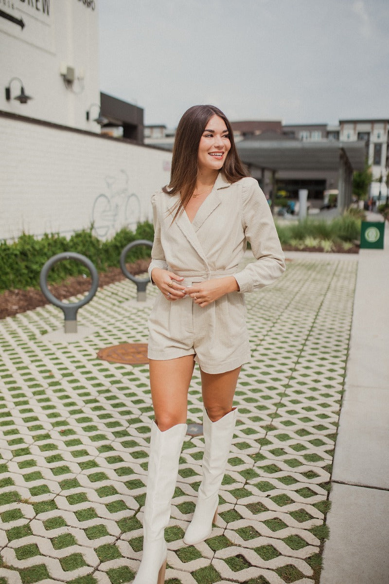 Frontal view of the More Than Enough Romper that features an ivory corduroy material, a collar neck, a surplice front, a long sleeve with a button, a front zipper, pockets, and pleating on the legs