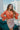 Frontal view of the Daphne Bodysuit that features a copper colored silk material, a surplice neckline with a button closure, a long sleeve with a ruffle detail at the wrist, and a bikini bottom with a snap closure.