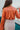Back view of the Daphne Bodysuit that features a copper colored silk material, a surplice neckline with a button closure, a long sleeve with a ruffle detail at the wrist, and a bikini bottom with a snap closure.