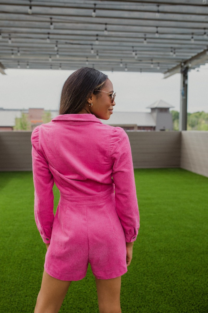  Back view of the More Than Enough Romper that features a pink corduroy material, a collar neck, a surplice front, a long sleeve with a button, a front zipper, pockets, and pleating on the legs.