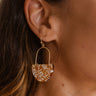 The Hard To Get Earring is a dangle style earring, featuring an open gold arch shape, a half circle wood body, and a white floral print.